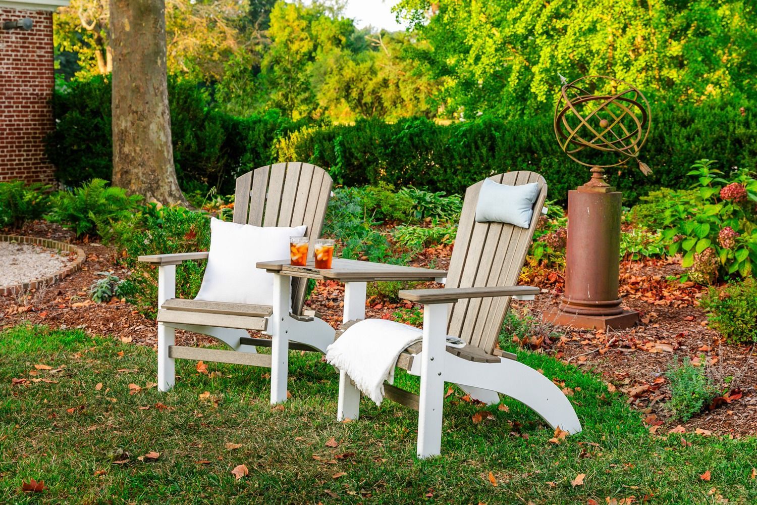 Two light-colored Adirondack chairs with a shared table sit on a lush lawn near a metal armillary sphere garden ornament.