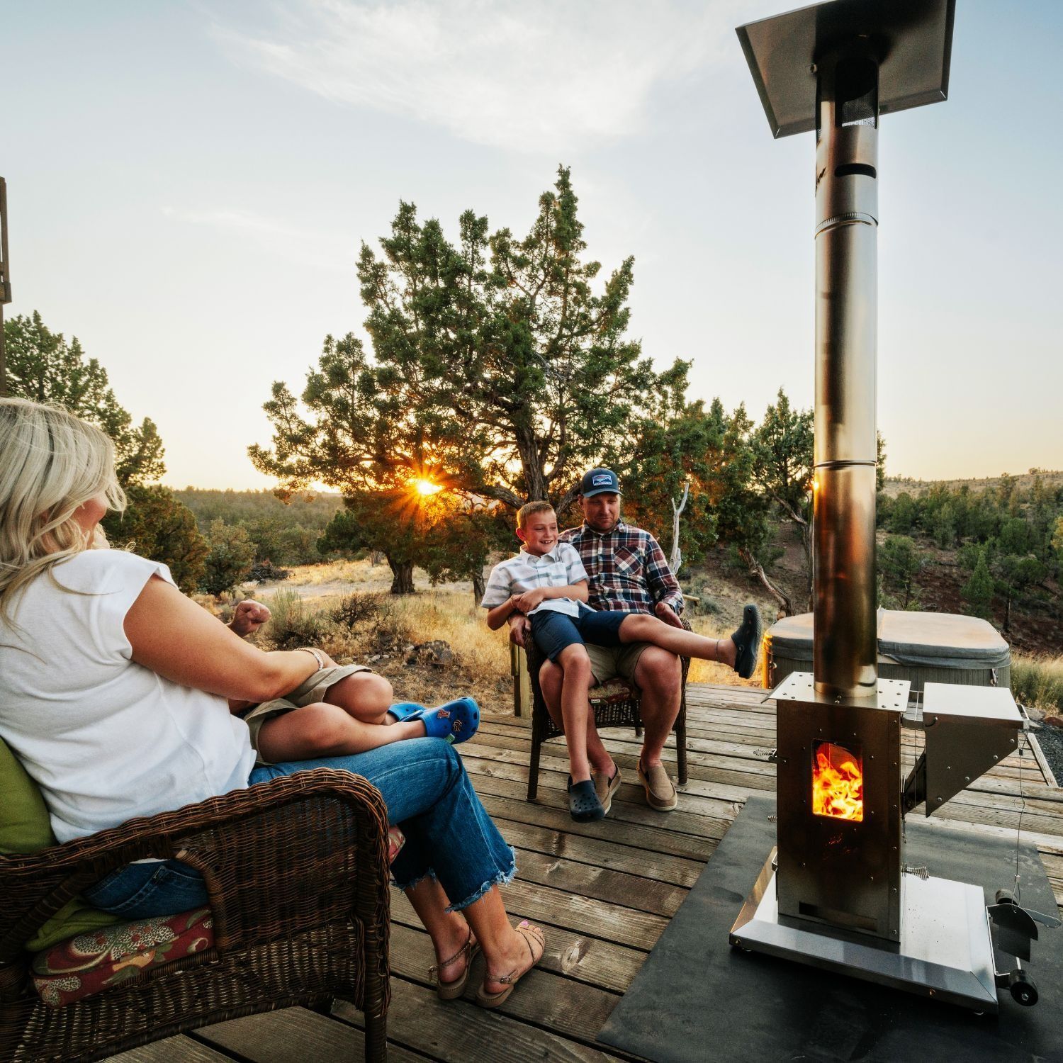 People relaxing outdoors around a fire pit at sunset, seated in wicker chairs on a deck.