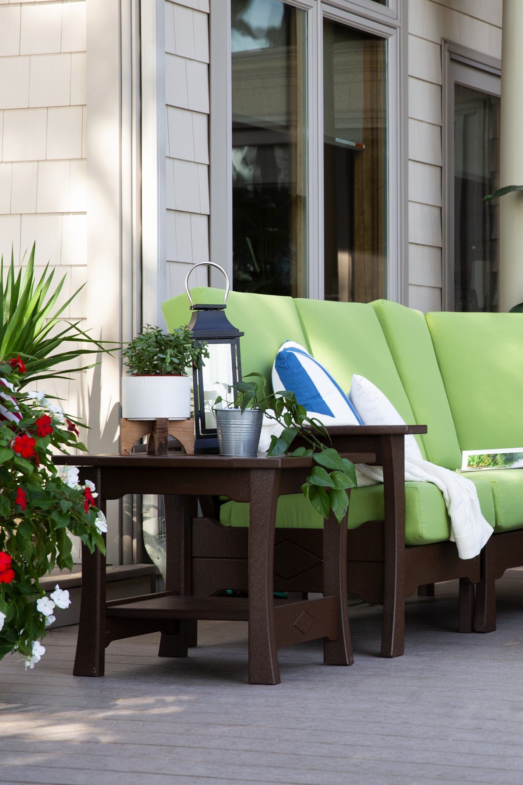 A patio features a bright green outdoor sofa, a dark wooden side table with potted plants, and a white, shingled wall.