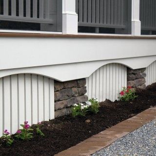 A patio features a bright green outdoor sofa, a dark wooden side table with potted plants, and a white, shingled wall.