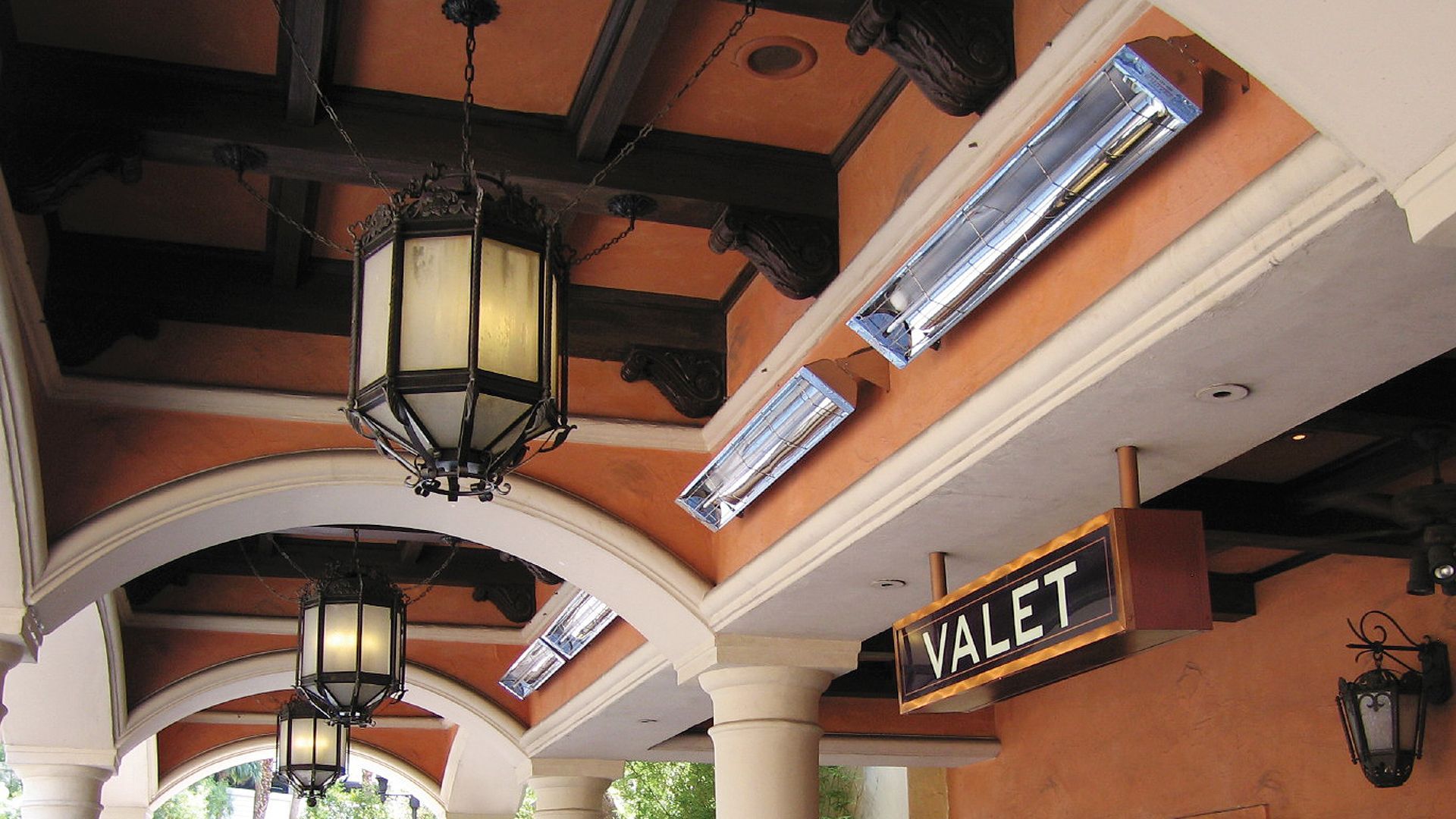 An outdoor valet entrance with warm-toned stucco walls, arched ceilings, hanging lanterns, and overhead heaters.