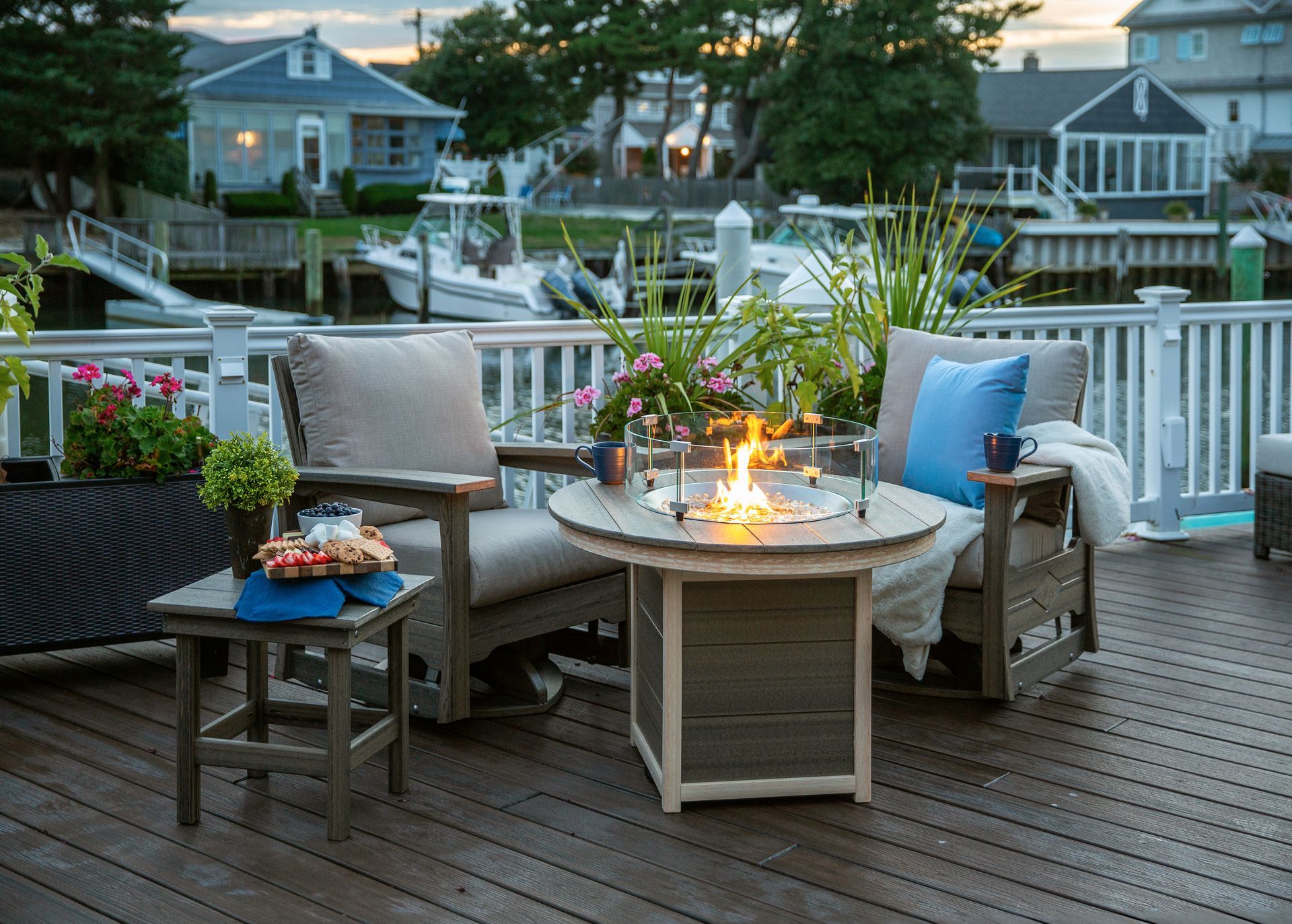 Two wooden chairs with light gray cushions face a circular fire pit on a dark wood deck overlooking a marina with boats.
