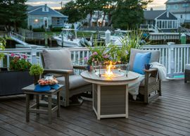 Two wooden chairs with light gray cushions face a circular fire pit on a dark wood deck overlooking a marina with boats.