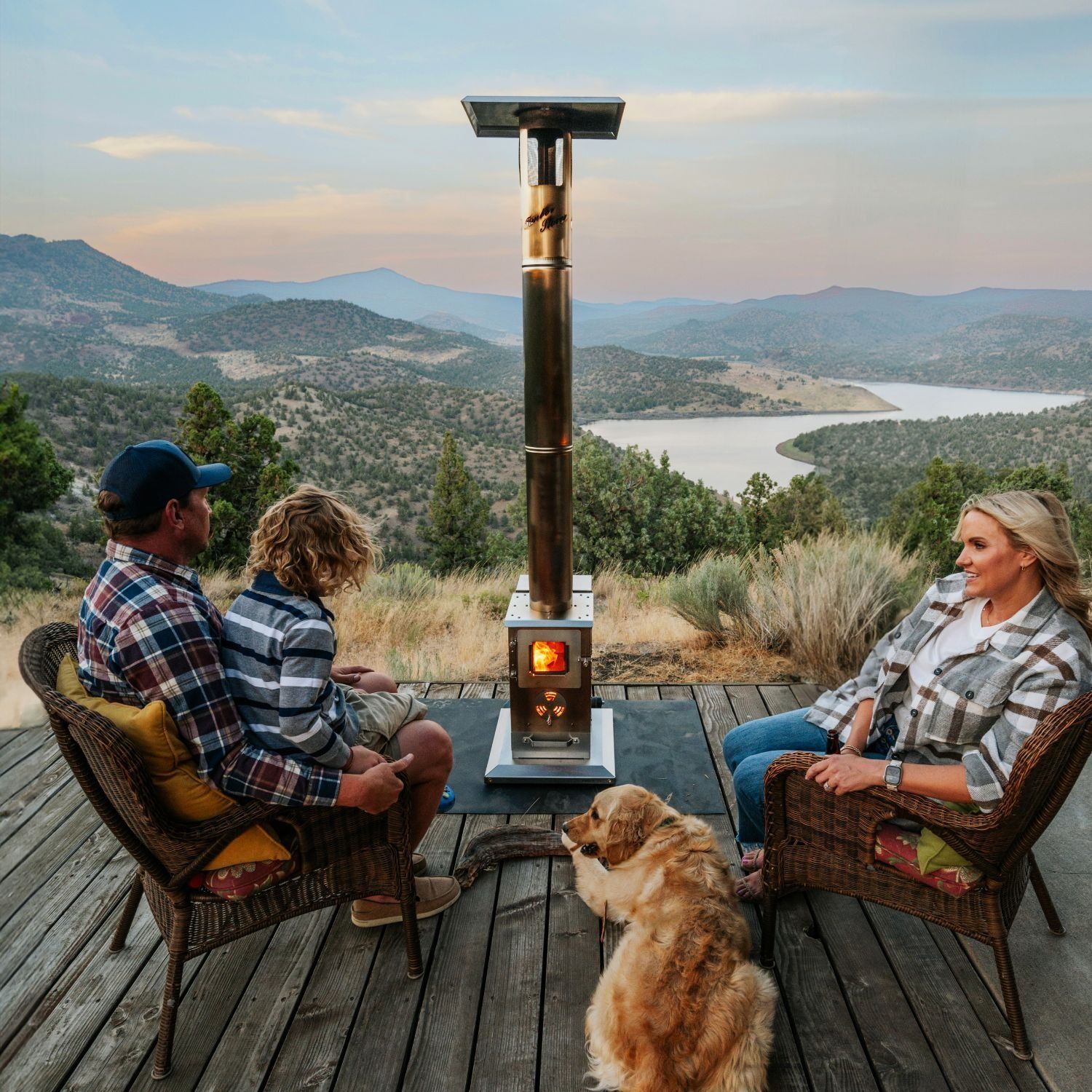 Two people and a dog sit by a wood stove on a deck overlooking a river valley at sunset