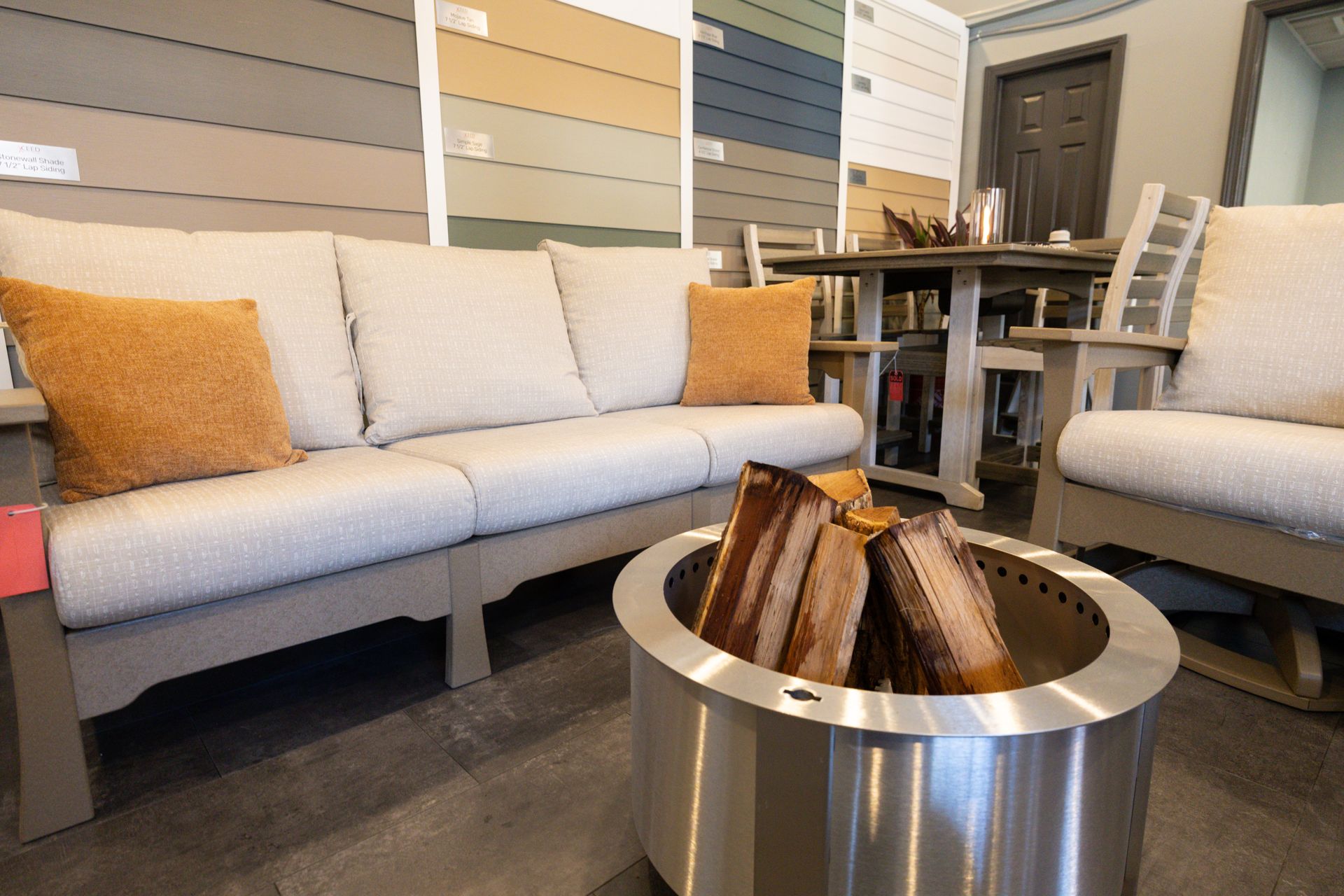 A showroom display with a light-colored outdoor sofa, fire pit, and dining set against a backdrop of siding samples.