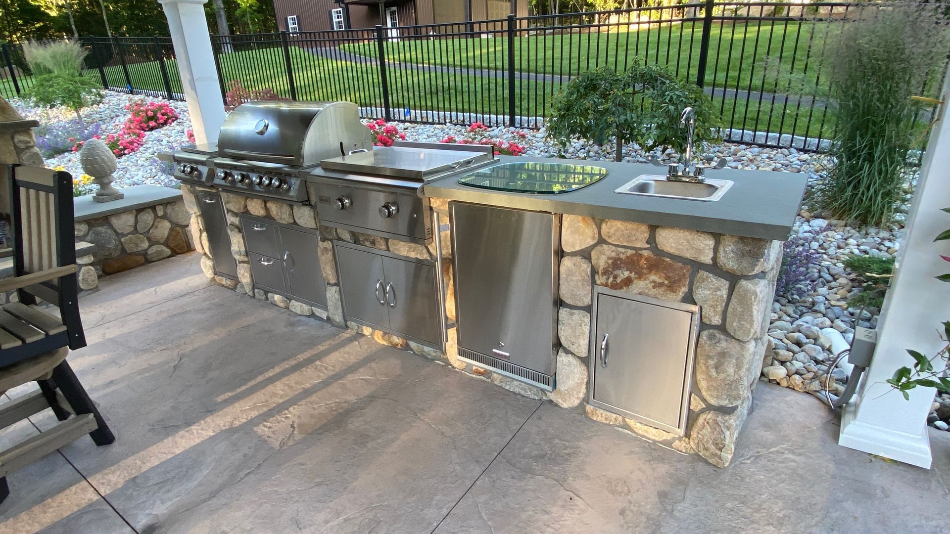 An outdoor kitchen with a stainless steel grill, side burner, sink, and stone facade on a stone patio.