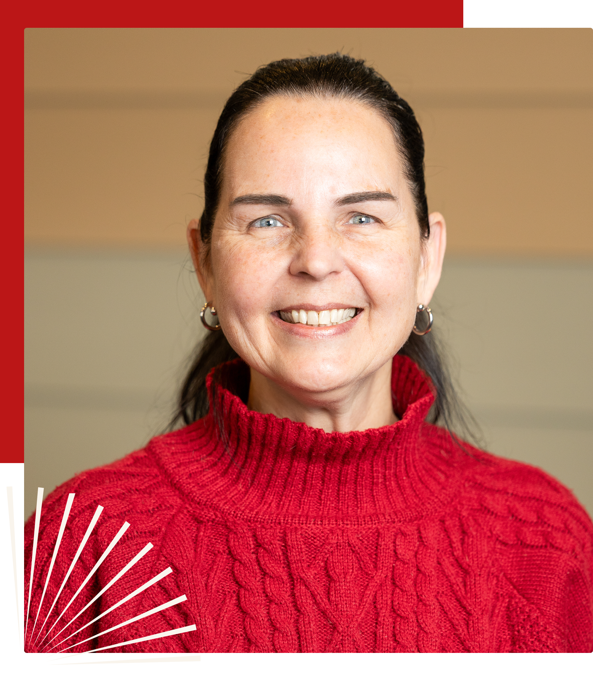 A smiling person with dark hair, wearing a red cable-knit sweater against a neutral, paneled wall background.