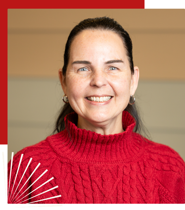 A smiling person with dark hair, wearing a red cable-knit sweater against a neutral, paneled wall background.