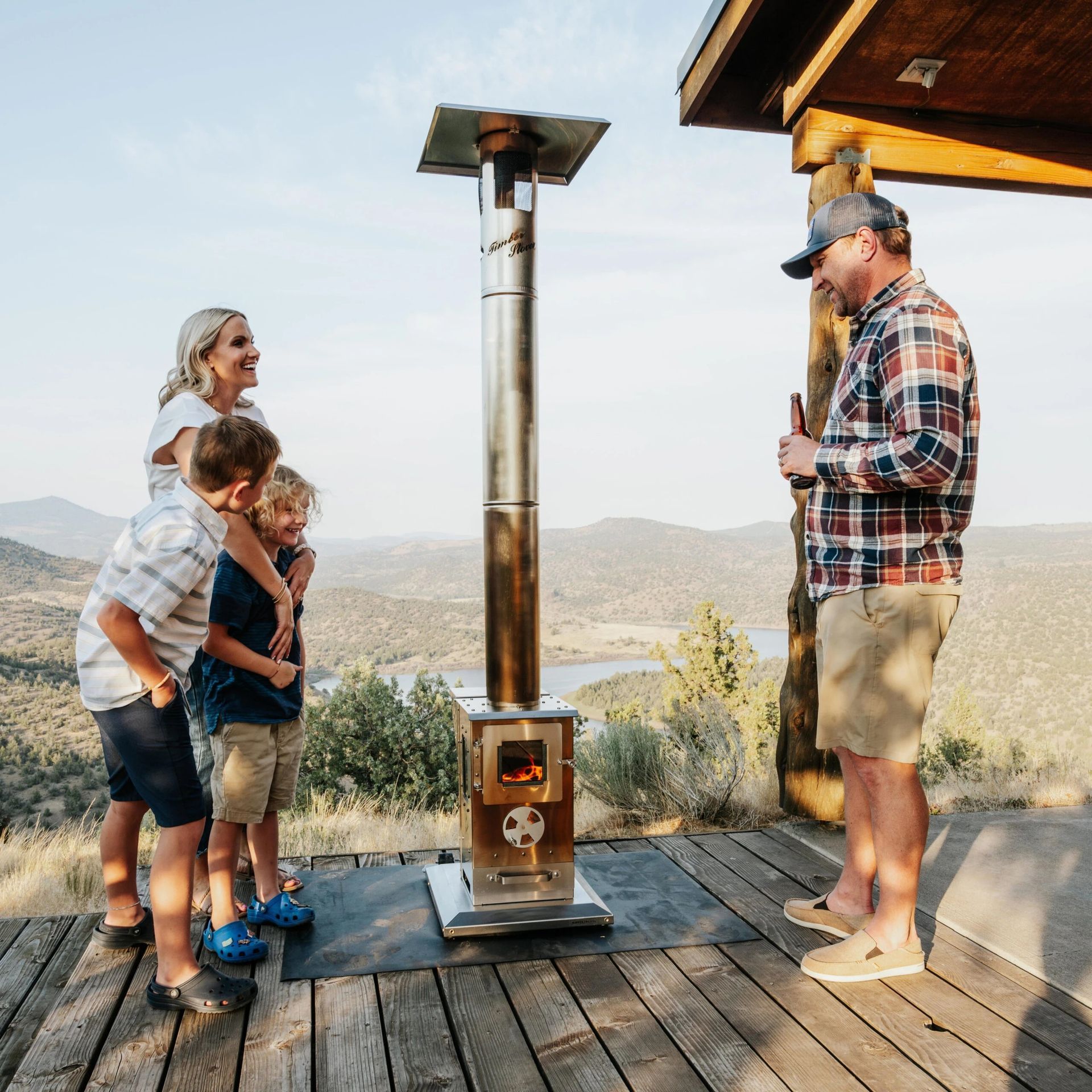 A family gathers on a wooden deck around a small, lit outdoor stove overlooking a mountain landscape.