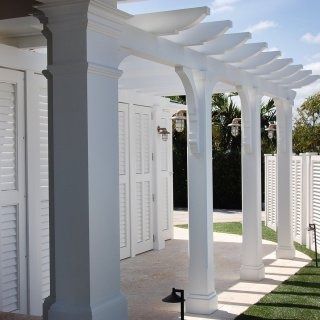 A white pergola with square columns and shutter-style walls stands over a light-colored patio walkway in a bright space.