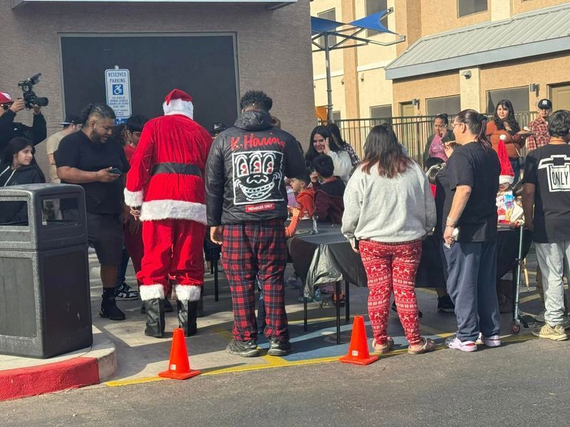 Santa, people in line, presents, outdoors. A festive gathering near a building with a blue awning.