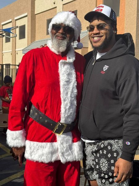 Man in Santa suit and young man in sunglasses pose outdoors.