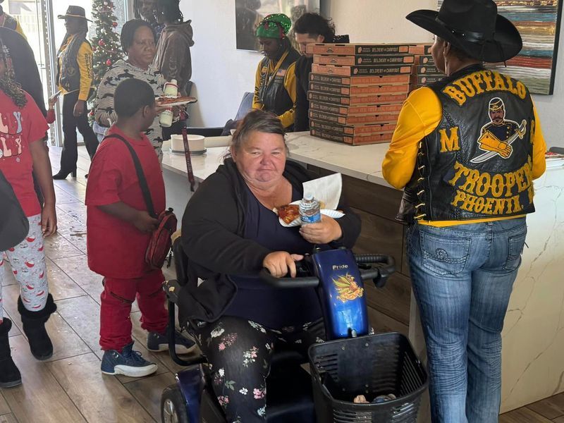 A woman in a wheelchair eats food while other people stand near a counter with pizza boxes. Buffalo Troopers logo on a vest.