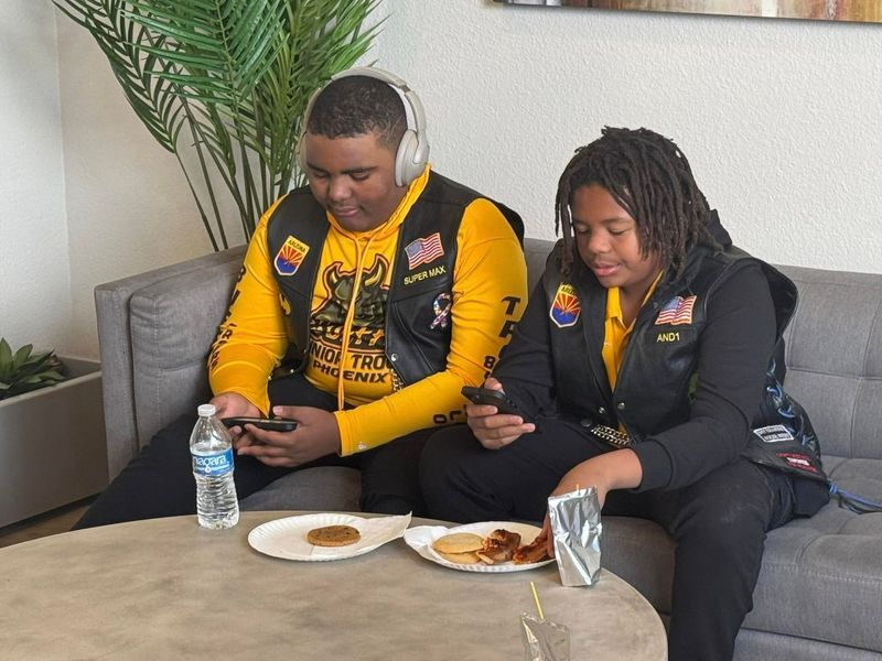 Two people on a couch, wearing vests and hoodies, looking at their phones. Food and water on the table.