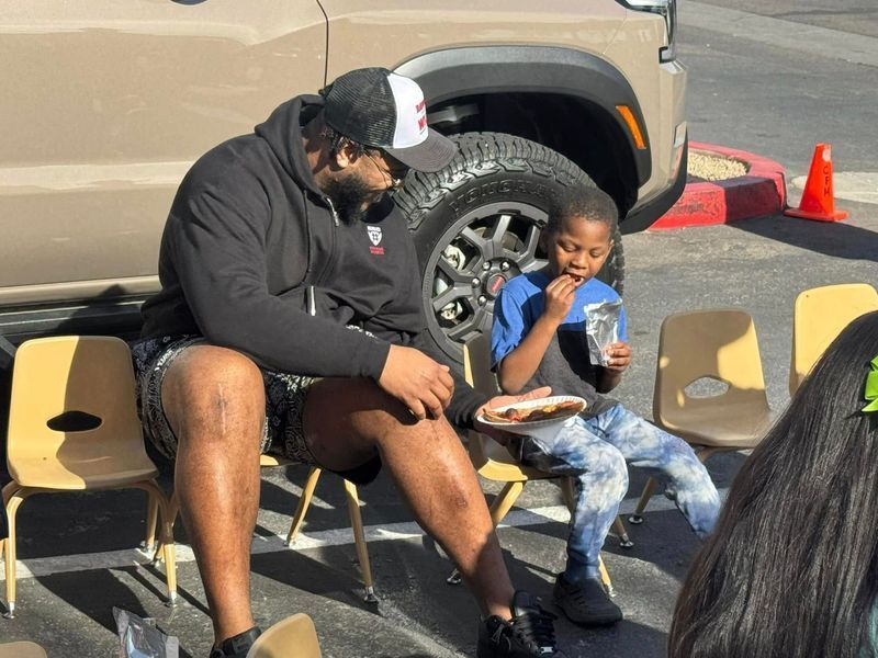 Man in black hoodie and boy seated eating, near vehicle and chairs.