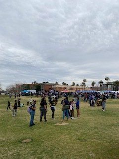 People gather on a grassy field in front of a building under a cloudy sky.