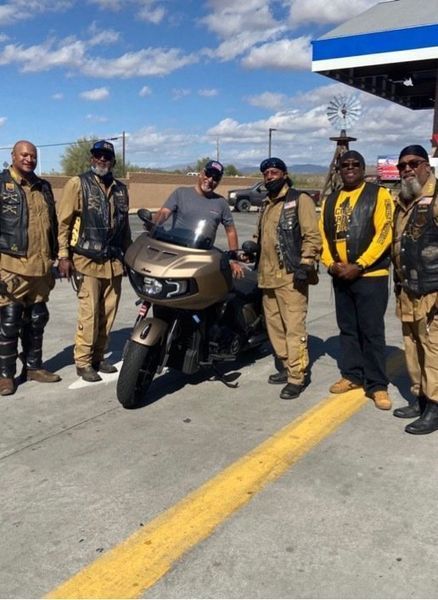 Motorcycle riders pose with a gold motorcycle at a gas station under a blue sky.