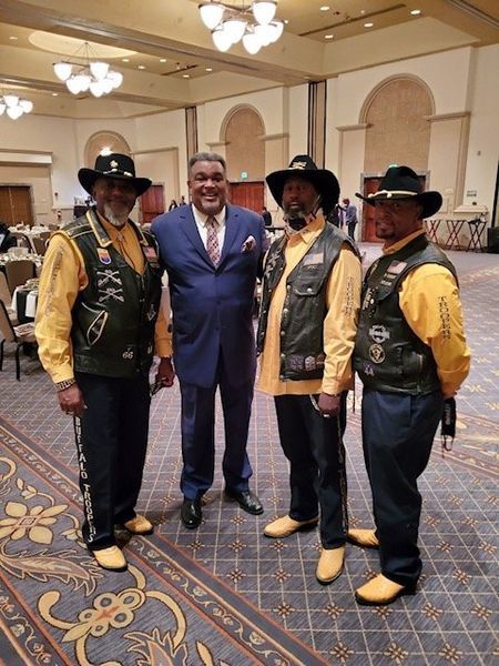 Four men in cowboy hats and vests with a man in a suit, posing together in a ballroom.