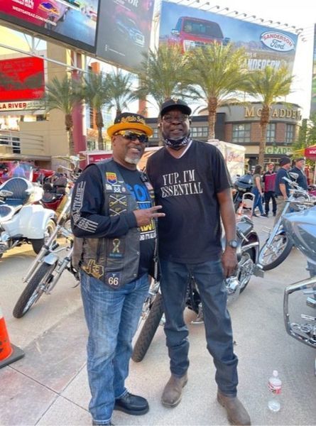 Two men pose for photo with motorcycles in outdoor setting. One wears biker vest.
