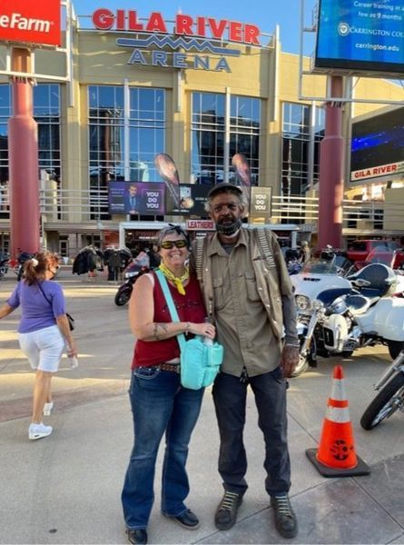 Two people pose in front of Gila River Arena; motorcycles parked nearby.