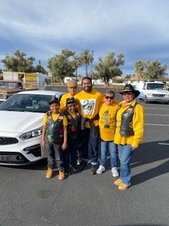 Group of six people in yellow shirts, vests, and hats standing next to a white car in a parking lot.