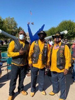 Three people in yellow shirts, black vests, and cowboy boots pose outdoors by a monument.