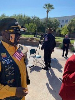 Man in uniform with US flag pin, standing outdoors with others, sunny day.