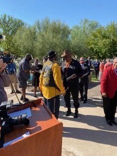 Two men in uniform shaking hands outdoors, surrounded by people and a podium.
