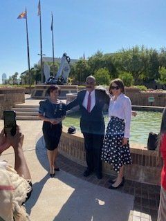 Three people pose near a fountain. A man in a suit stands between two women; all smile. The background shows a monument and buildings.