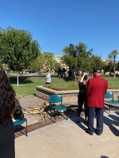People standing outside near a grassy area with trees, chairs, and a building under a blue sky.