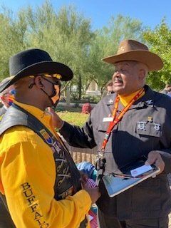 Two men in hats talking outdoors; one in yellow and black, the other in a military-style uniform.