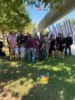 Group of people gathered outdoors near a large, white pipe structure. Sunny day.