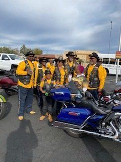 Group of bikers in yellow and black vests pose by motorcycles. Some wear cowboy hats, setting is outdoors.