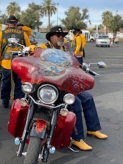 Man on red motorcycle, wearing hat and yellow shirt, parked outside. Other riders in background.