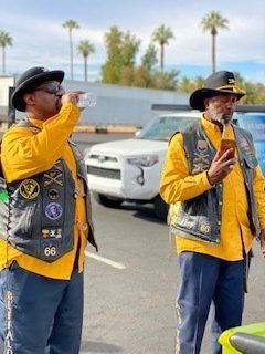 Two men in vests, hats, yellow shirts, and blue pants near a car, one drinking, the other on a phone.