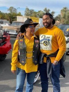 Woman in biker vest and hat with man in yellow shirt, smiling, posing outdoors.
