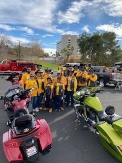 Group of people in yellow vests pose with motorcycles outdoors. Building and sky in the background.
