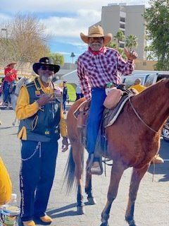 Two men in Western attire; one on horseback, waving. Outdoors, sunny day.