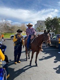 A person on a horse, wearing a cowboy hat, next to a person in a vest, outdoors in a parking lot.