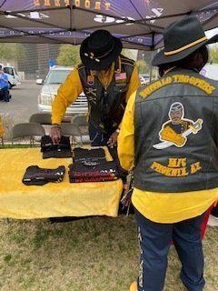 Two people in yellow and black vests, Buffalo Soldiers MC Phoenix, selling merchandise at a table outdoors.
