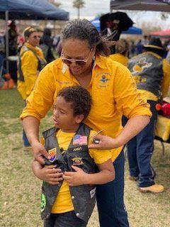 Woman in yellow shirt adjusts a child's vest at an outdoor event. Both wear similar outfits.