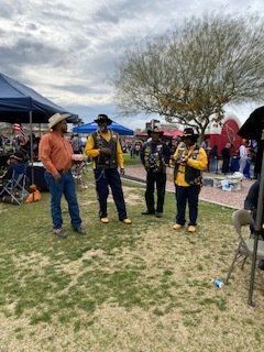 Four people in cowboy hats and vests stand outdoors; one in an orange shirt, two in yellow, and one in black.