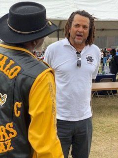 Man in white shirt talking to someone wearing a biker vest and hat outdoors.