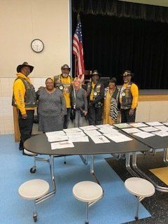 Group of people with American flag, gathered at a table in a room. Some wearing yellow vests.