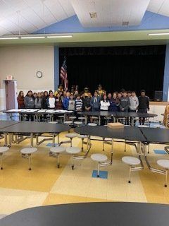 Group of people standing on a stage with an American flag. Tables and stools are in the foreground.