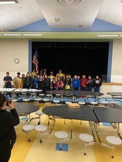 Students stand in a cafeteria in front of a stage with an American flag.