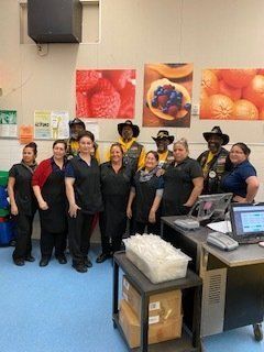 Group of people, some in biker attire, pose with grocery store employees near a checkout counter.