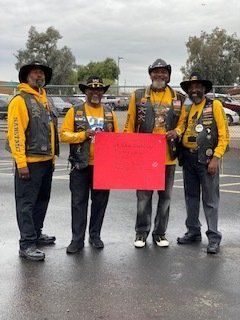 Four men in biker attire holding a red sign outside.