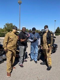 Four people in biker attire pose near motorcycles on a sunny day. Two wear brown jumpsuits; the others, jeans/t-shirts.