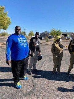 Four people standing outside on a sunny day. One man in blue shirt, others wearing neutral tones.